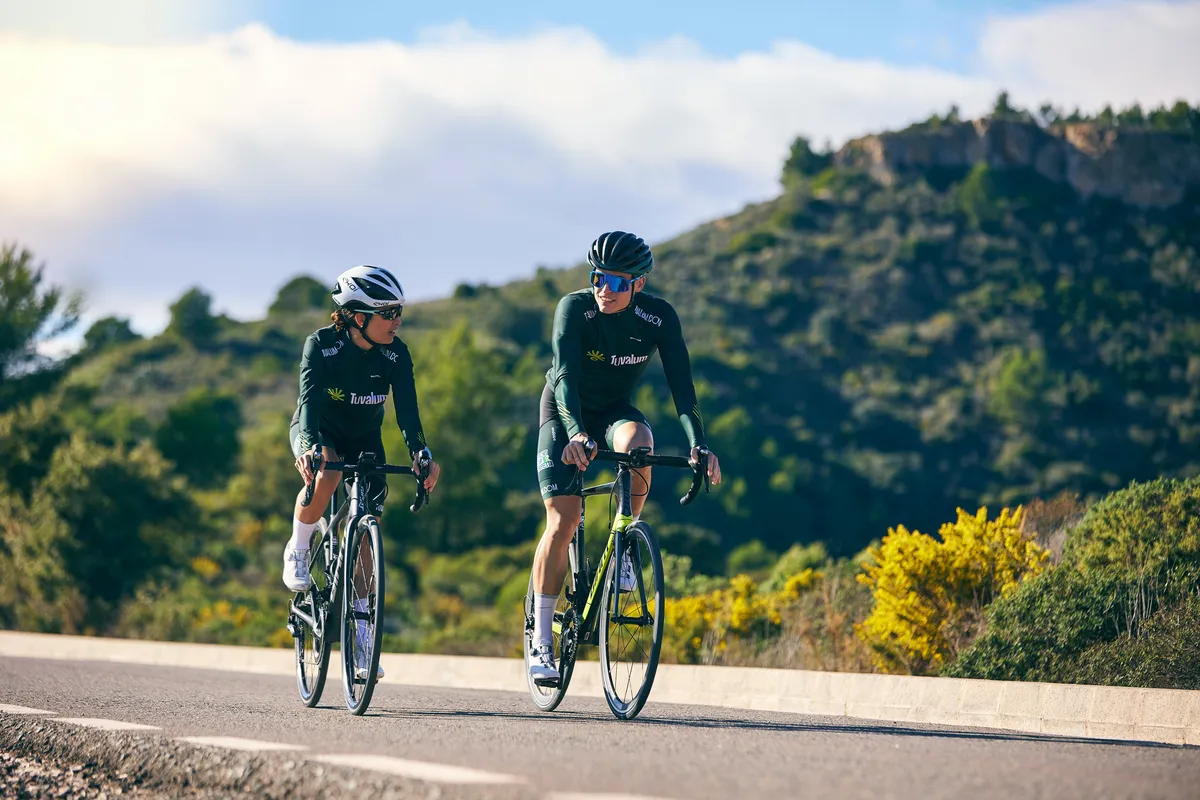 two men riding bikes on a road with trees in the background